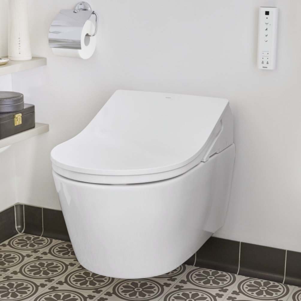 Lifestyle image of the TOTO Washlet RX Rimless Wall Hung Toilet mounted on a white bathroom wall next to a toilet roll holder and the Washlet remote control